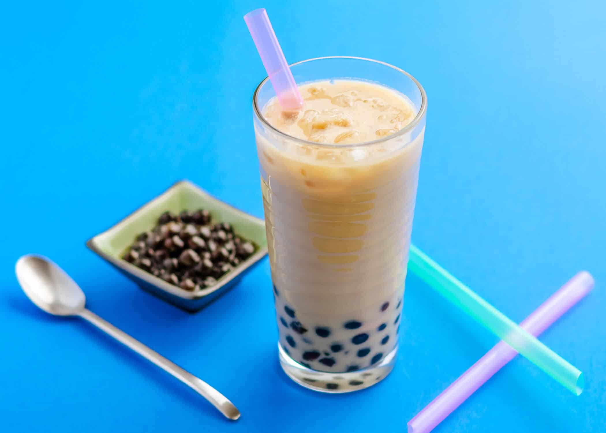 jasmine tea honey boba in a tall glass on a blue background with a bowl of boba pearls, straws and long spoon. 